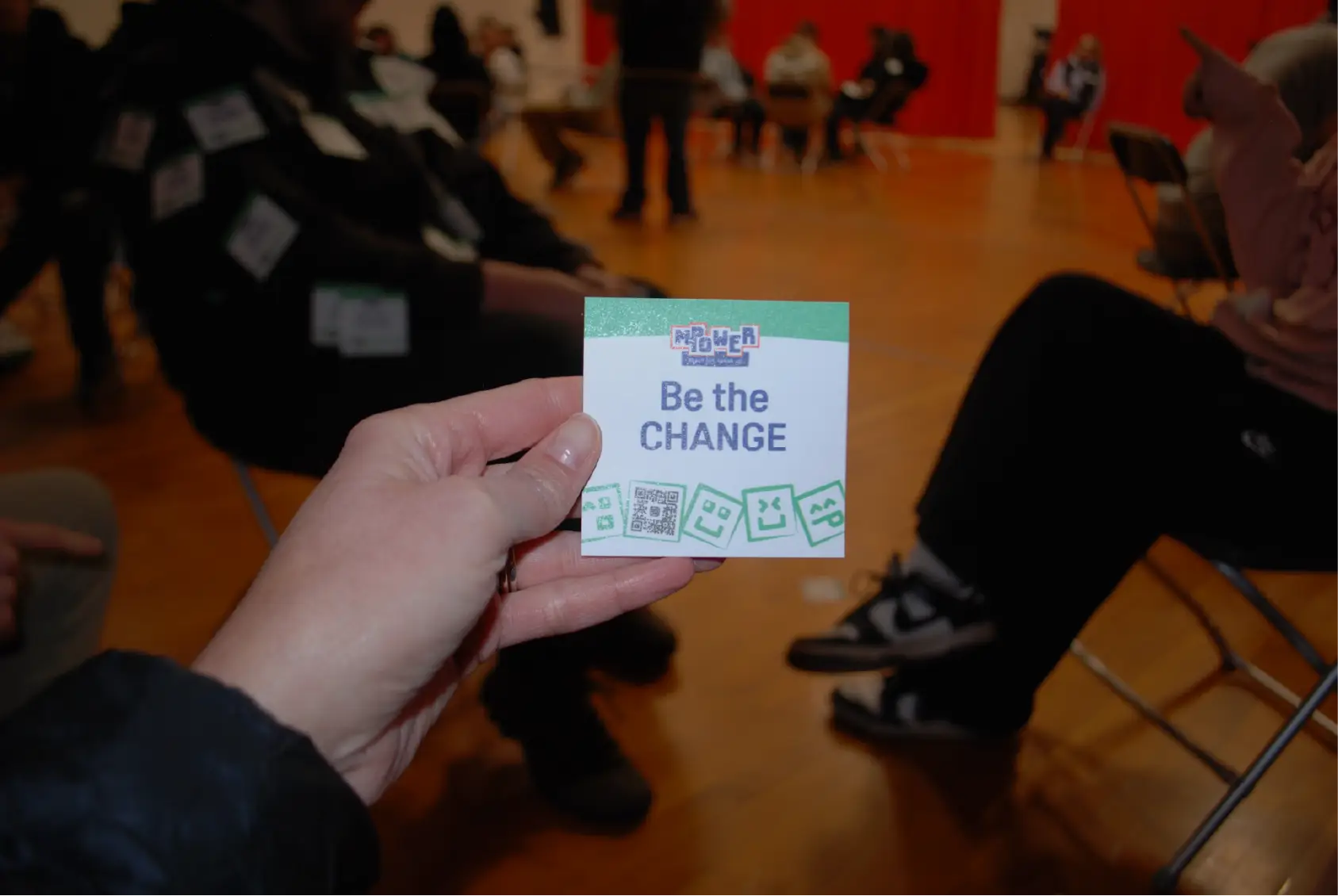 A hand holds a card reading Be the CHANGE in a room with people seated in a circle, fostering community and connection.