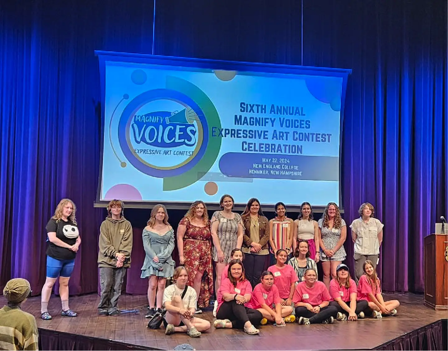 A group poses on stage at an art contest celebration, highlighting community spirit, with a large event banner displayed behind them.