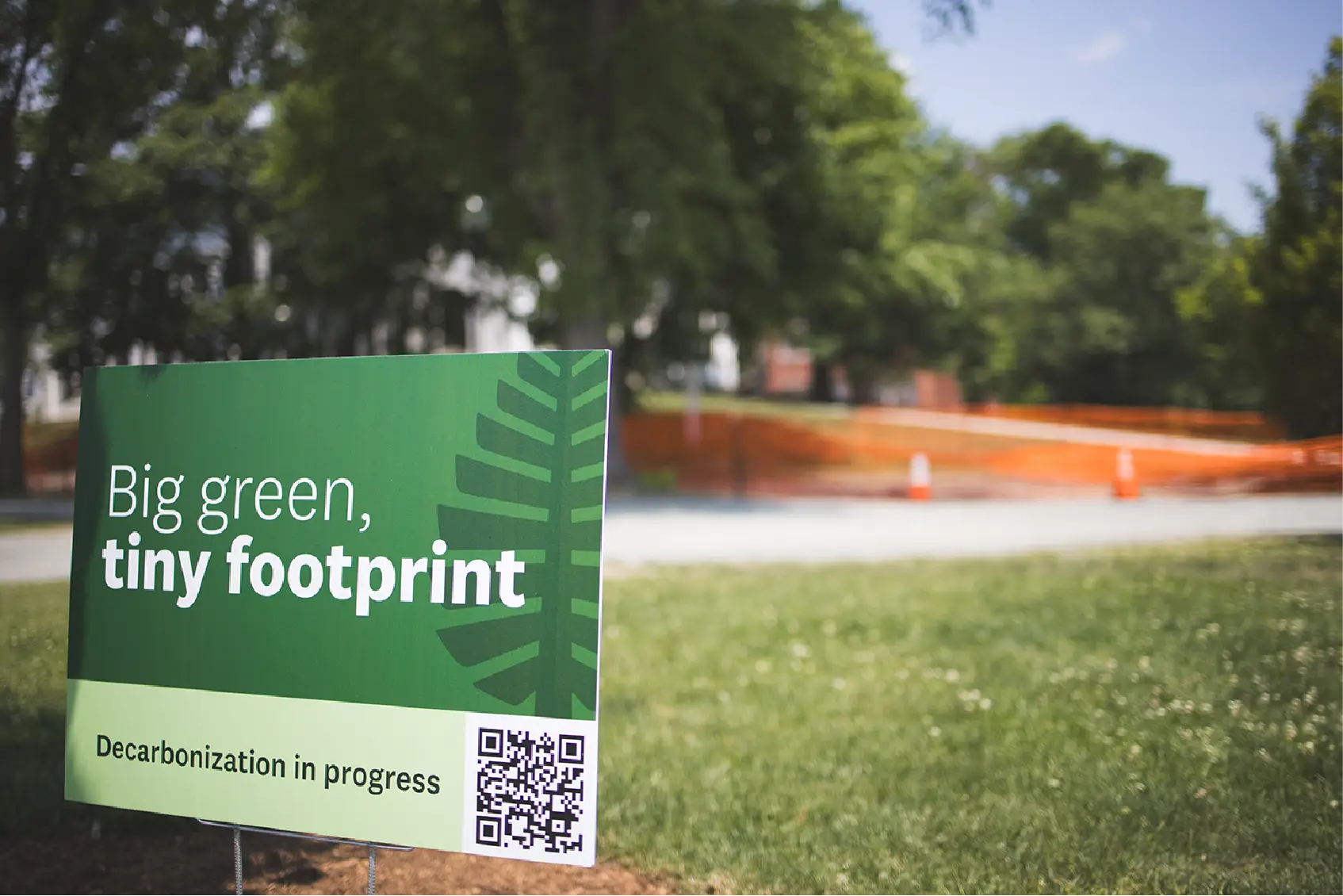 A green sign on grass reads Big green, tiny footprint. Decarbonization in progress, engaging the community with a QR code—a fresh take on sustainable marketing.