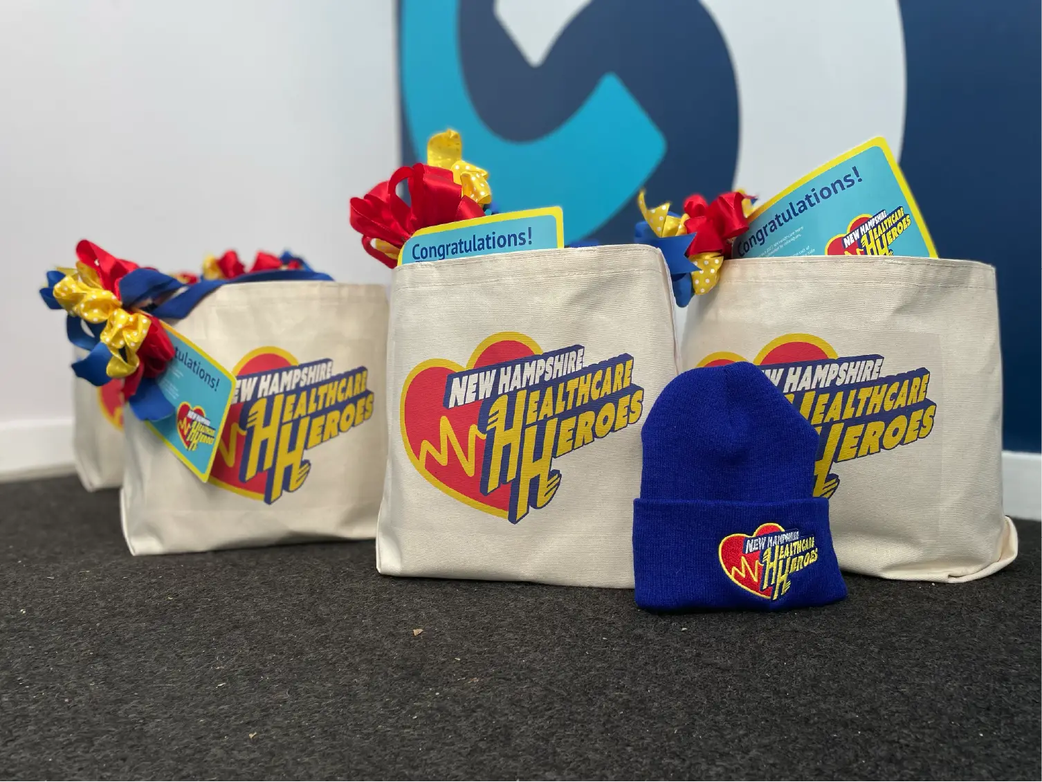 Gift bags labeled New Hampshire Healthcare Heroes and a blue beanie with the same logo showcase thoughtful community marketing on a carpeted floor.