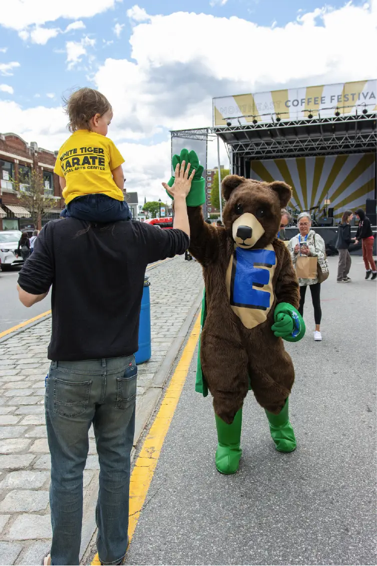 A child on an adult’s shoulders high-fives a bear mascot at an outdoor street festival, capturing the spirit of community.