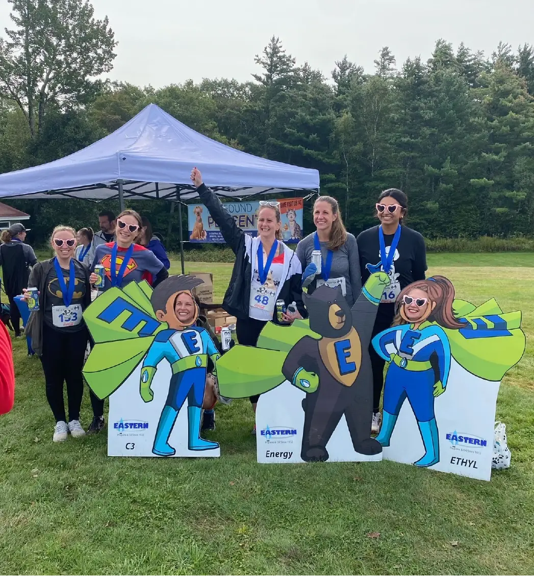 Six women with medals pose outdoors behind superhero cutouts at a race event, celebrating community spirit amidst trees in the background.