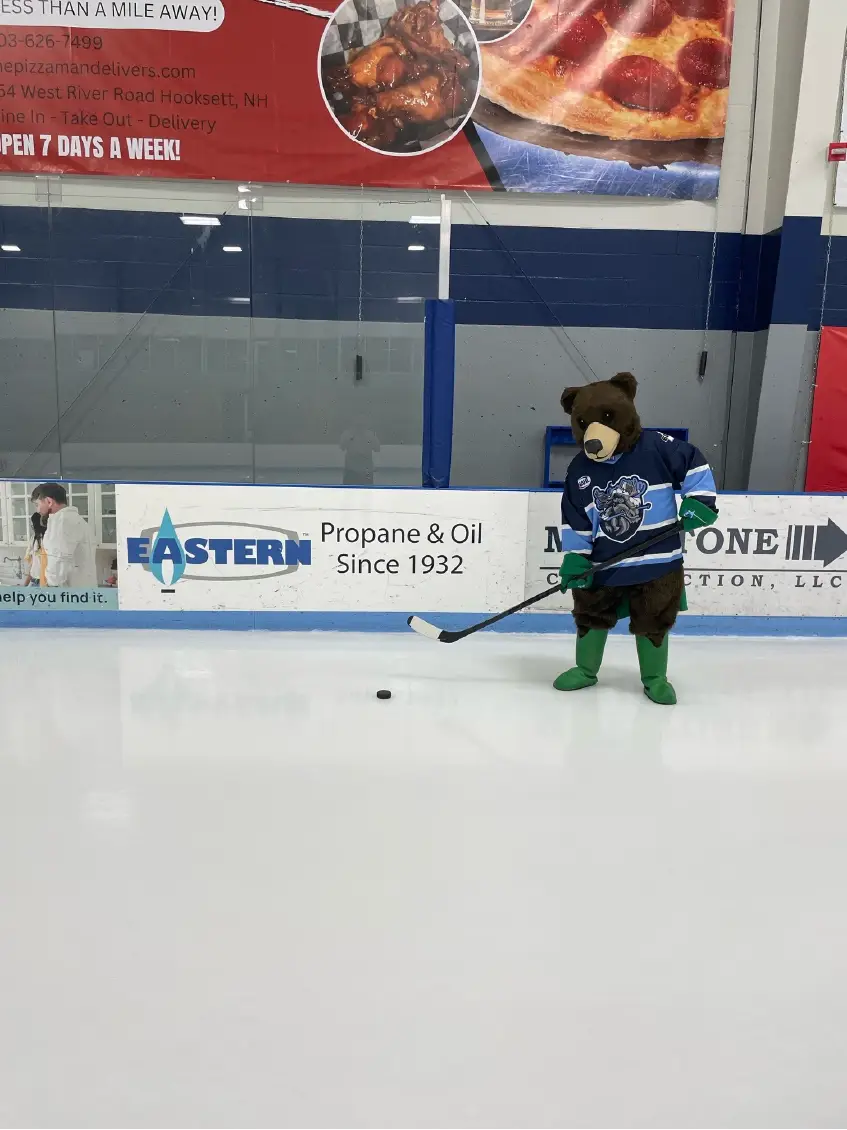 A person in a bear mascot hockey costume stands on an ice rink holding a hockey stick near a puck, bringing Community spirit and excitement to the event.