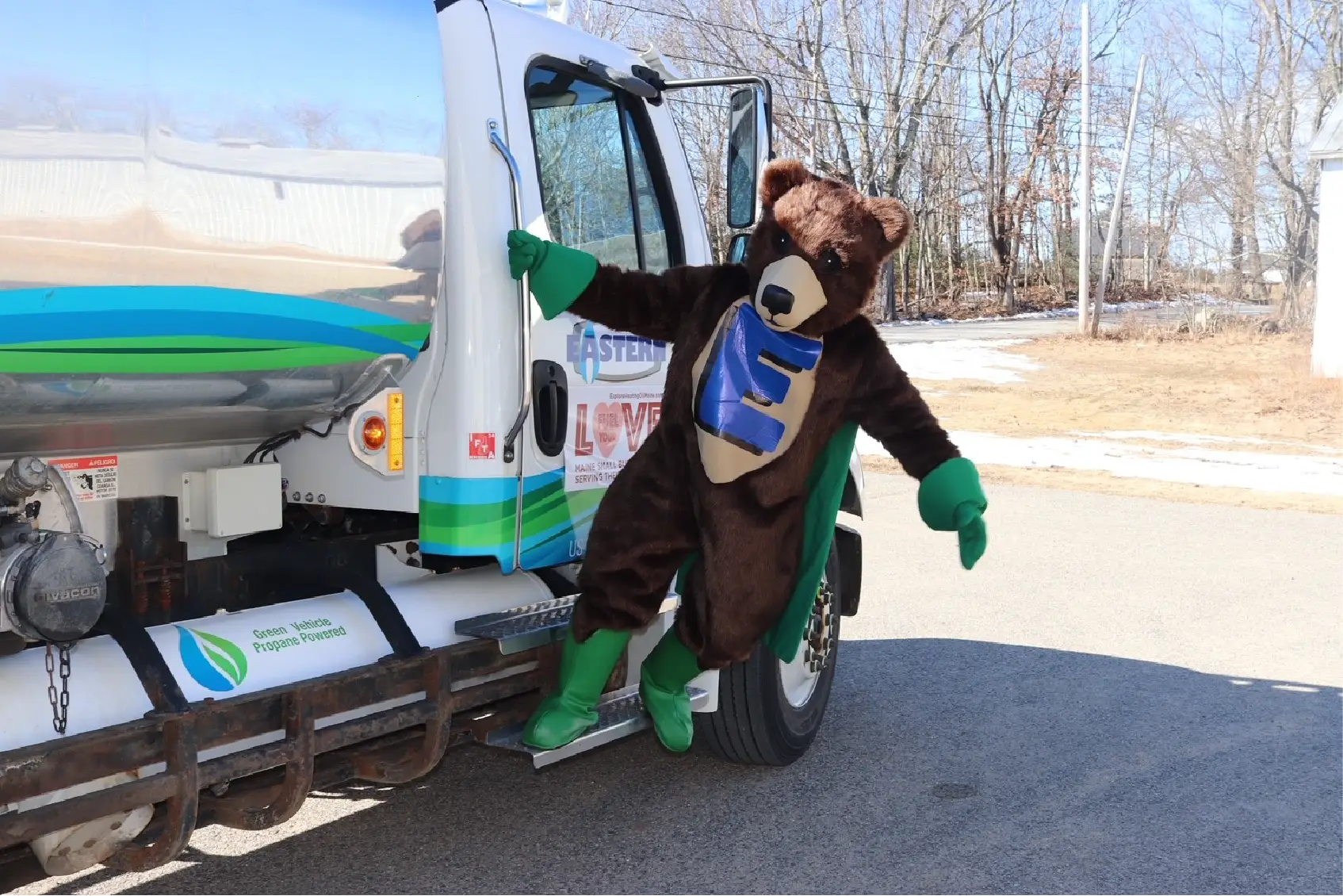 A person in a bear costume steps off a fuel truck on a sunny day, bringing smiles to the Community, with trees in the background.