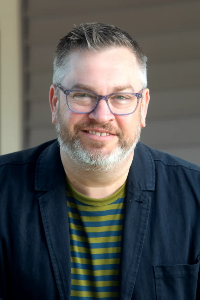 Smiling man with glasses, a gray beard, and short hair, wearing a striped shirt and dark blazer.