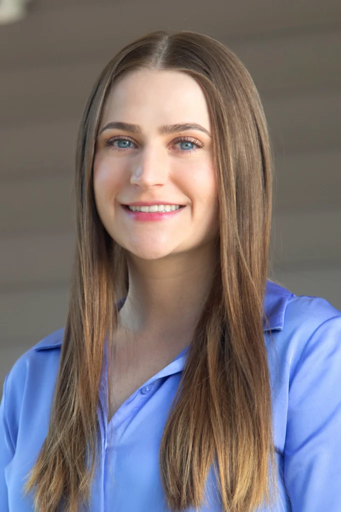 Smiling woman with long brown hair wearing a blue collared shirt, standing in front of a neutral background.