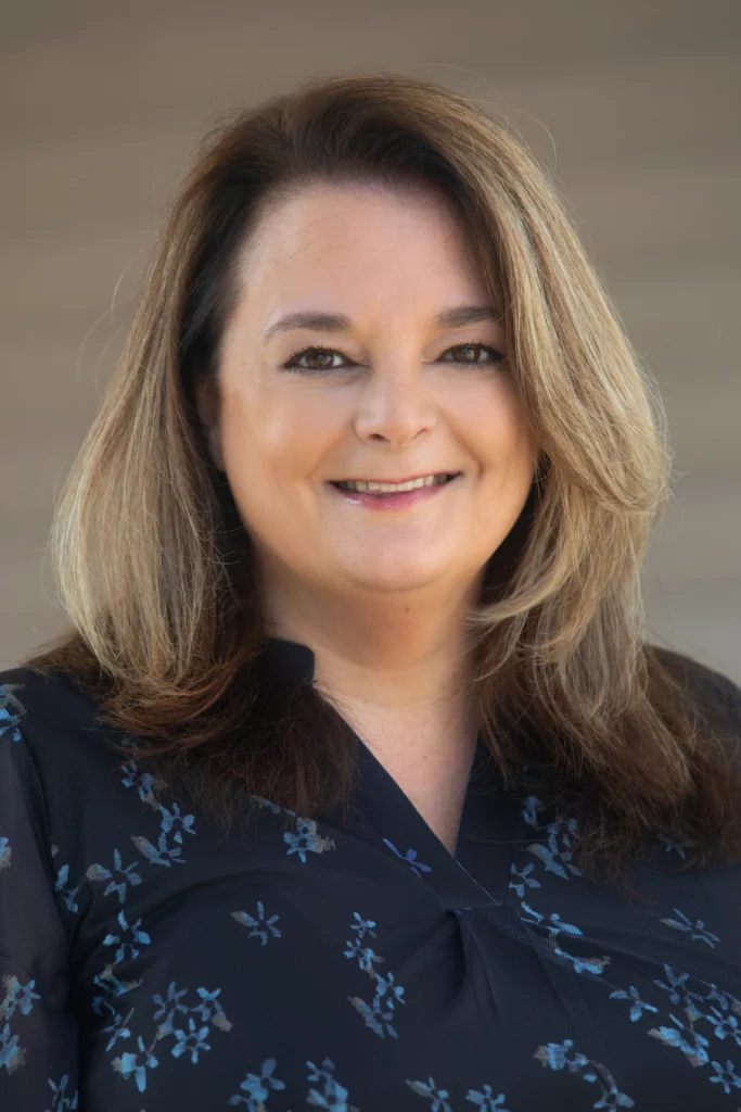 Jennifer Sperazzo, a woman with shoulder-length brown hair, smiles as she wears a dark blouse with a subtle floral pattern.