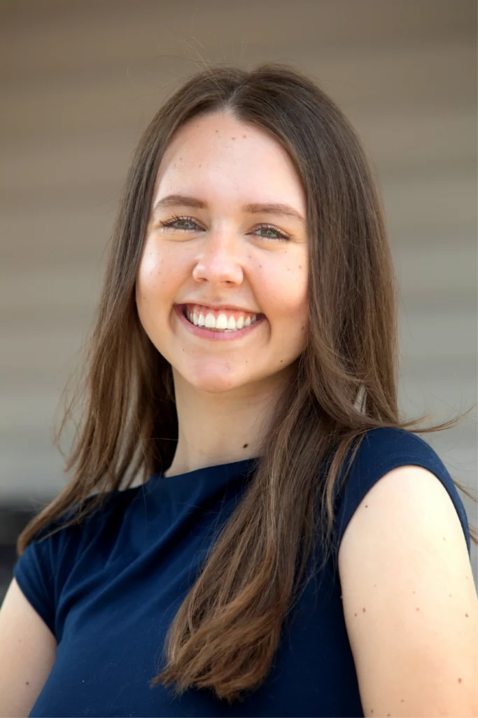 Emma Pedersen, a young woman with long brown hair, smiles warmly in a navy blue top while outdoors.