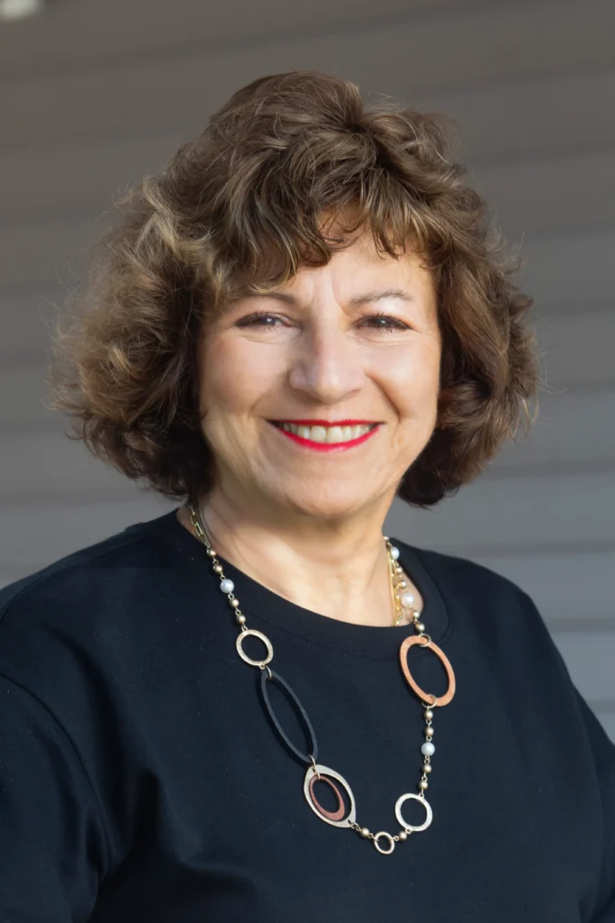 Deb LaPress, a smiling woman with curly brown hair, red lipstick, and a black top, wears a vibrant multicolored necklace.