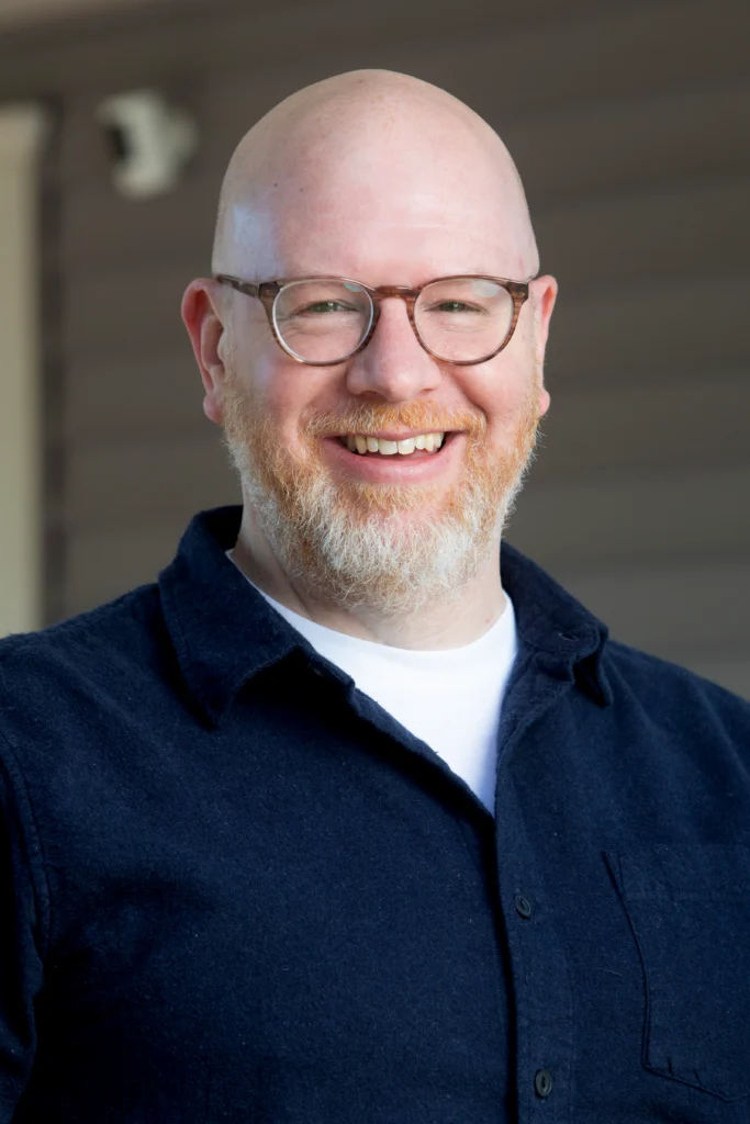 A smiling bald man with glasses and a beard, wearing a navy shirt and white t-shirt, stands outside—this is Steve Morin.