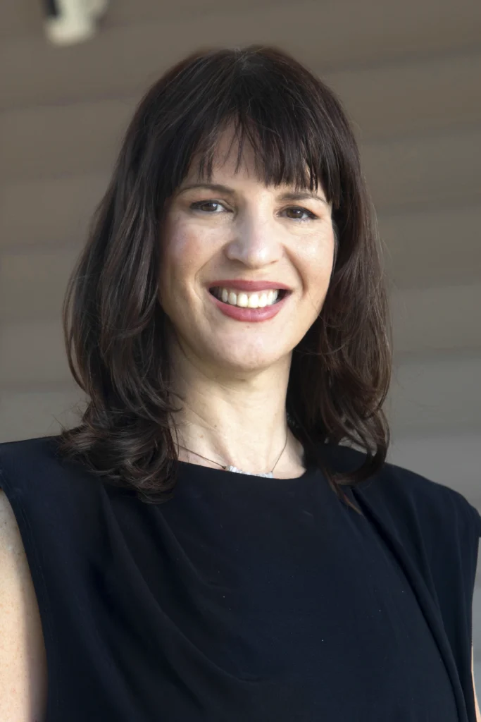 Woman with shoulder-length brown hair and bangs, smiling, wearing a sleeveless black top, standing indoors.