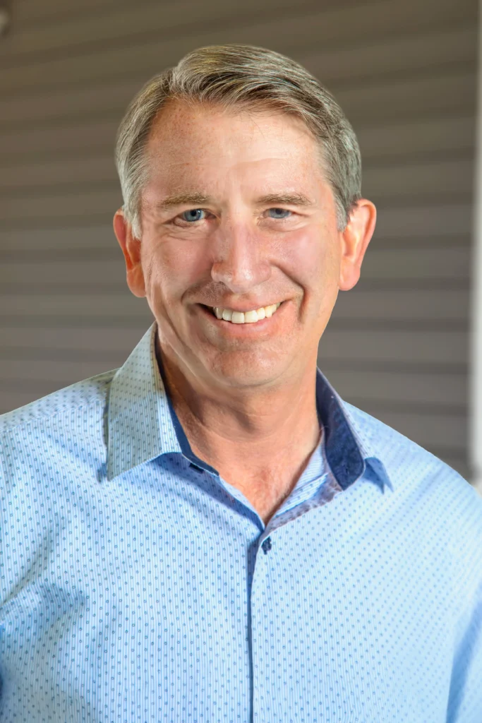 Smiling middle-aged man, Matt Cookson, with short gray hair wearing a light blue collared shirt, standing outdoors.