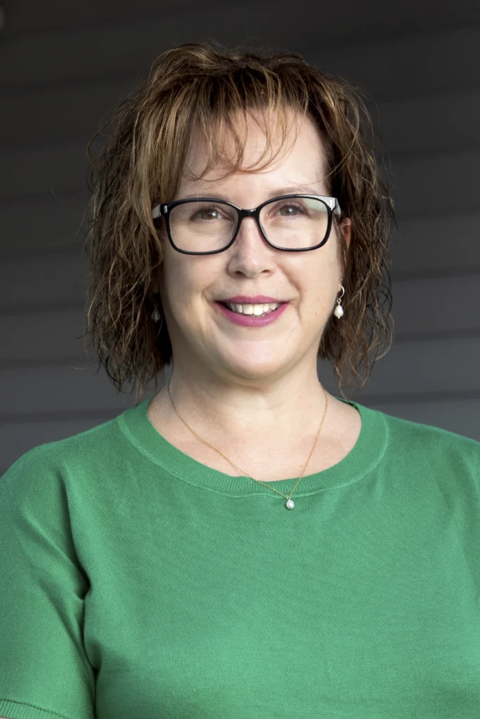 Woman with curly hair and glasses wearing a green sweater, smiling in front of a gray background.