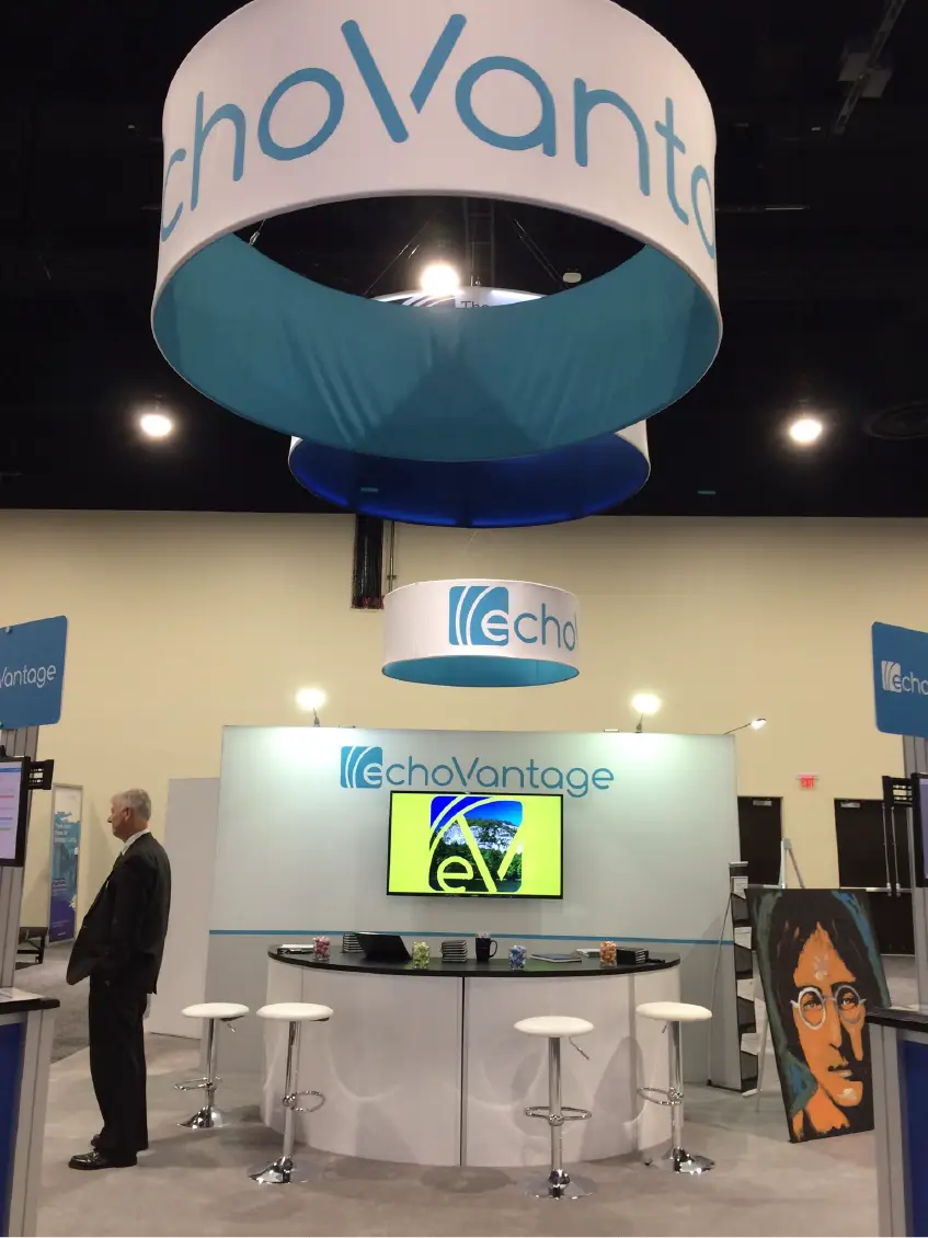 Trade show booth for echoVantage with a circular hanging sign, stools, and a portrait artwork on the right.