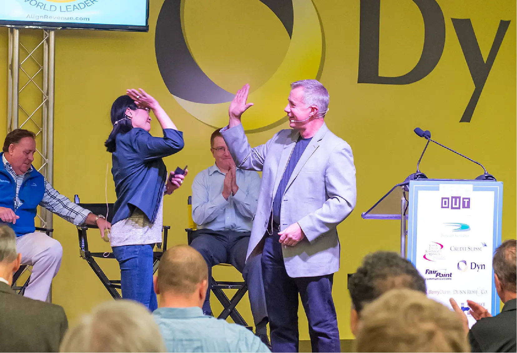 Two people on stage share a high five as others watch and applaud at a business event.
