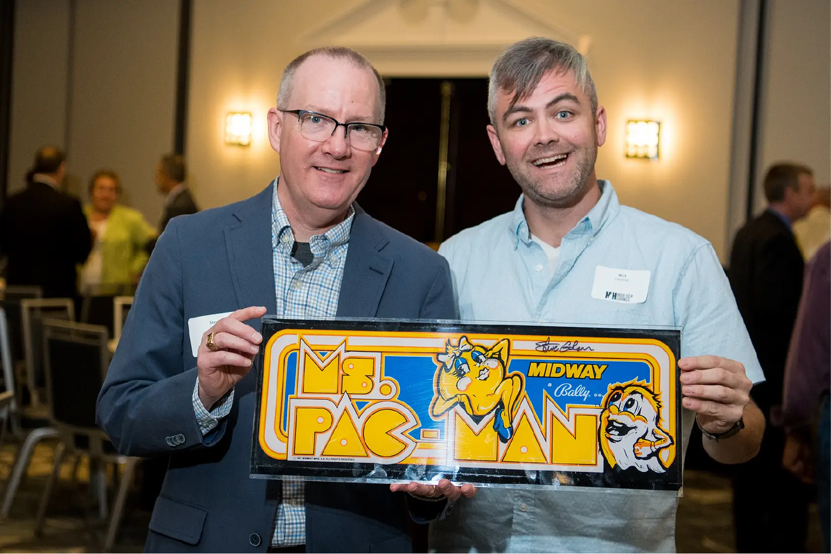 Two men smiling and holding a colorful Ms. Pac-Man arcade sign at an indoor event.