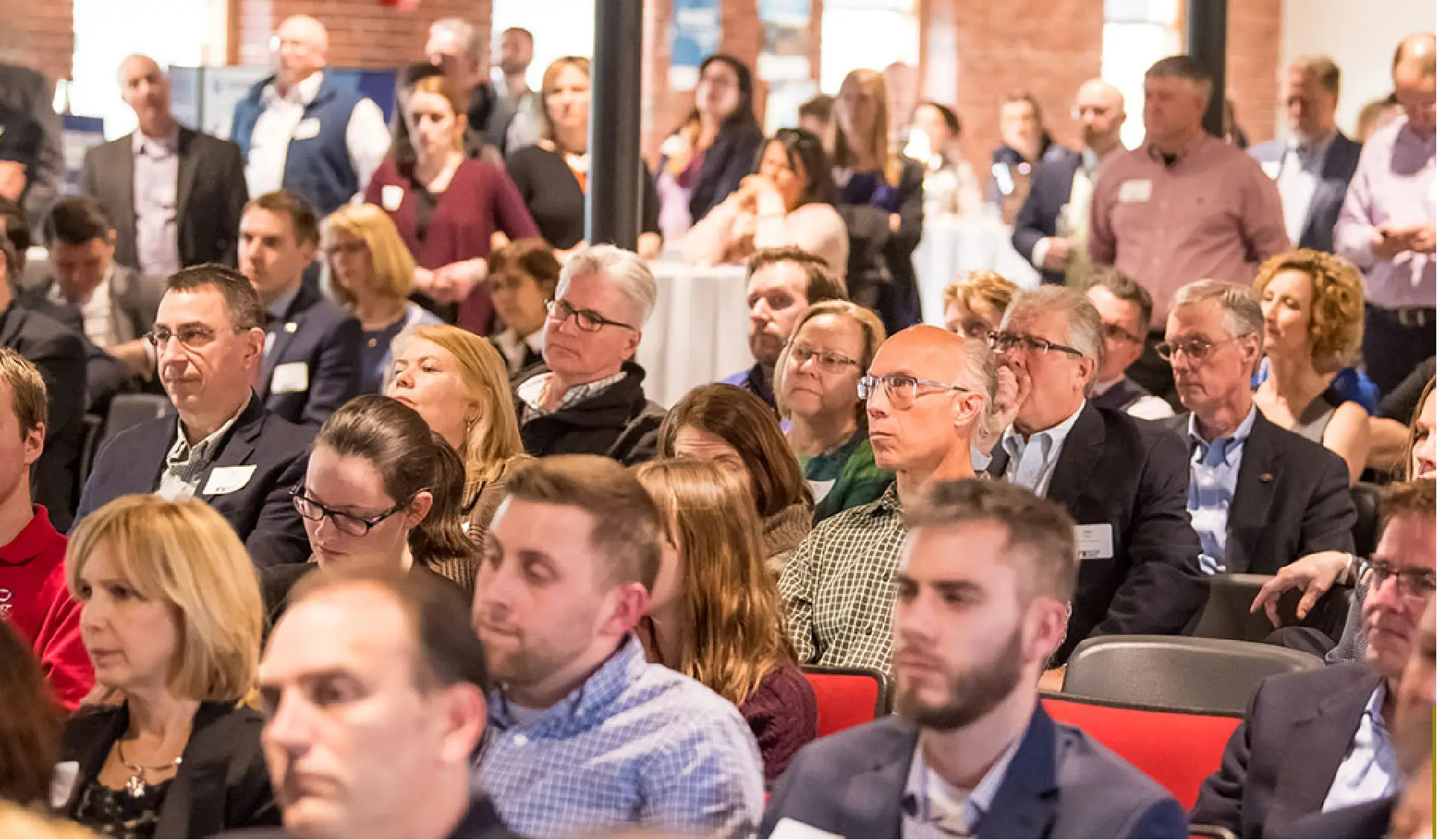 A large group of people sitting and standing at an indoor business conference or seminar.