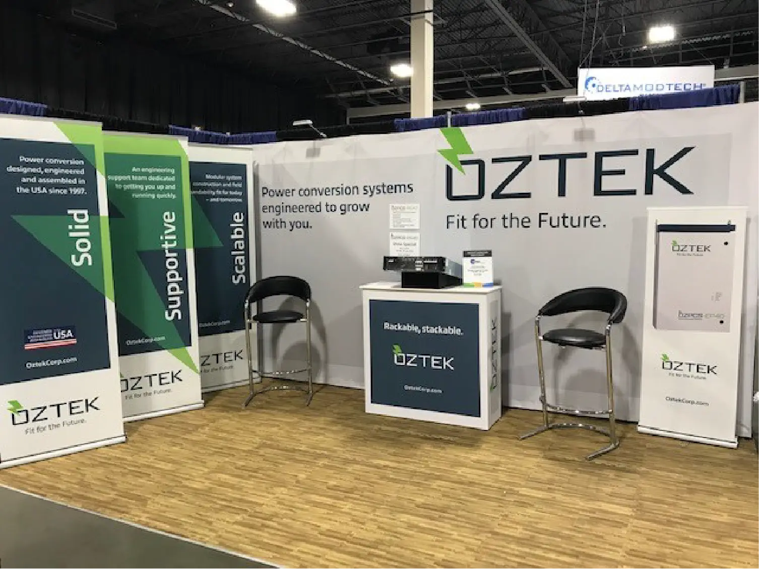 Oztek trade show booth with banners, two chairs, a table, and a brochure stand in an exhibition hall.