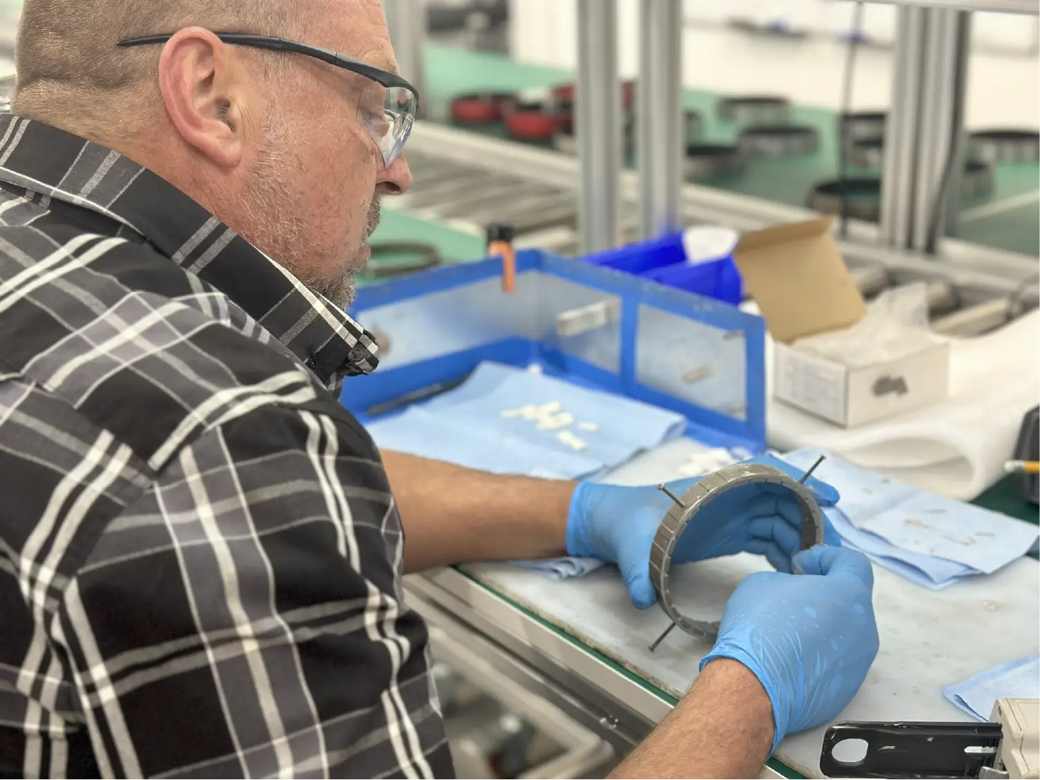 Man wearing gloves and safety glasses inspects a metal part at a workstation in a factory setting.