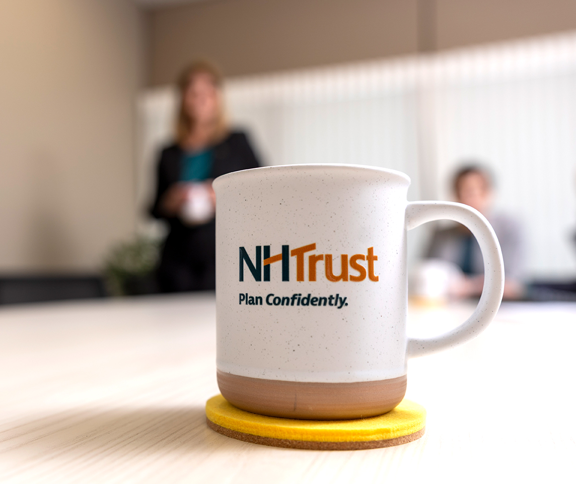 White mug with NHTrust Plan Confidently. logo on a desk, conveying a sense of finance expertise, with blurred people in the background.