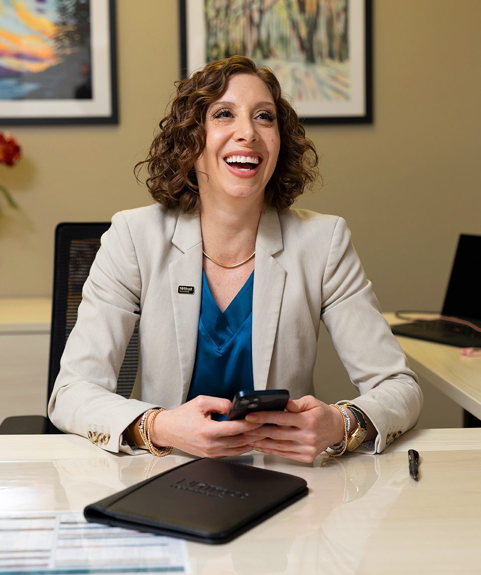 Woman with curly hair smiling, seated at a desk, holding a smartphone, with a folder and pen in front of her—ready to tackle her next finance project.