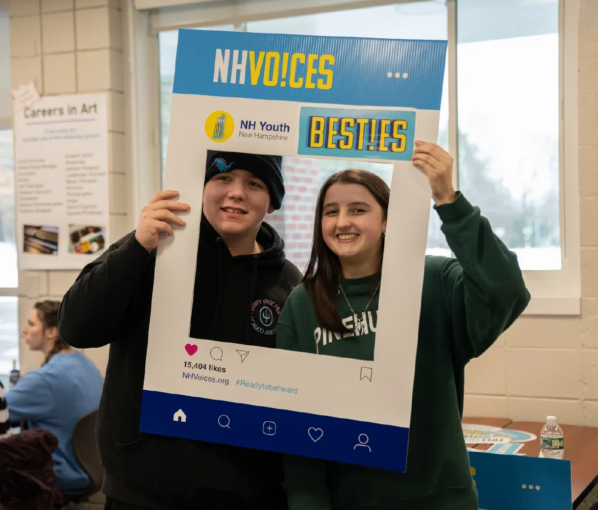 Two teens smile holding a blue NH Voices Besties photo frame at an indoor event, highlighting the power of friendship as part of an engaging brand strategy.