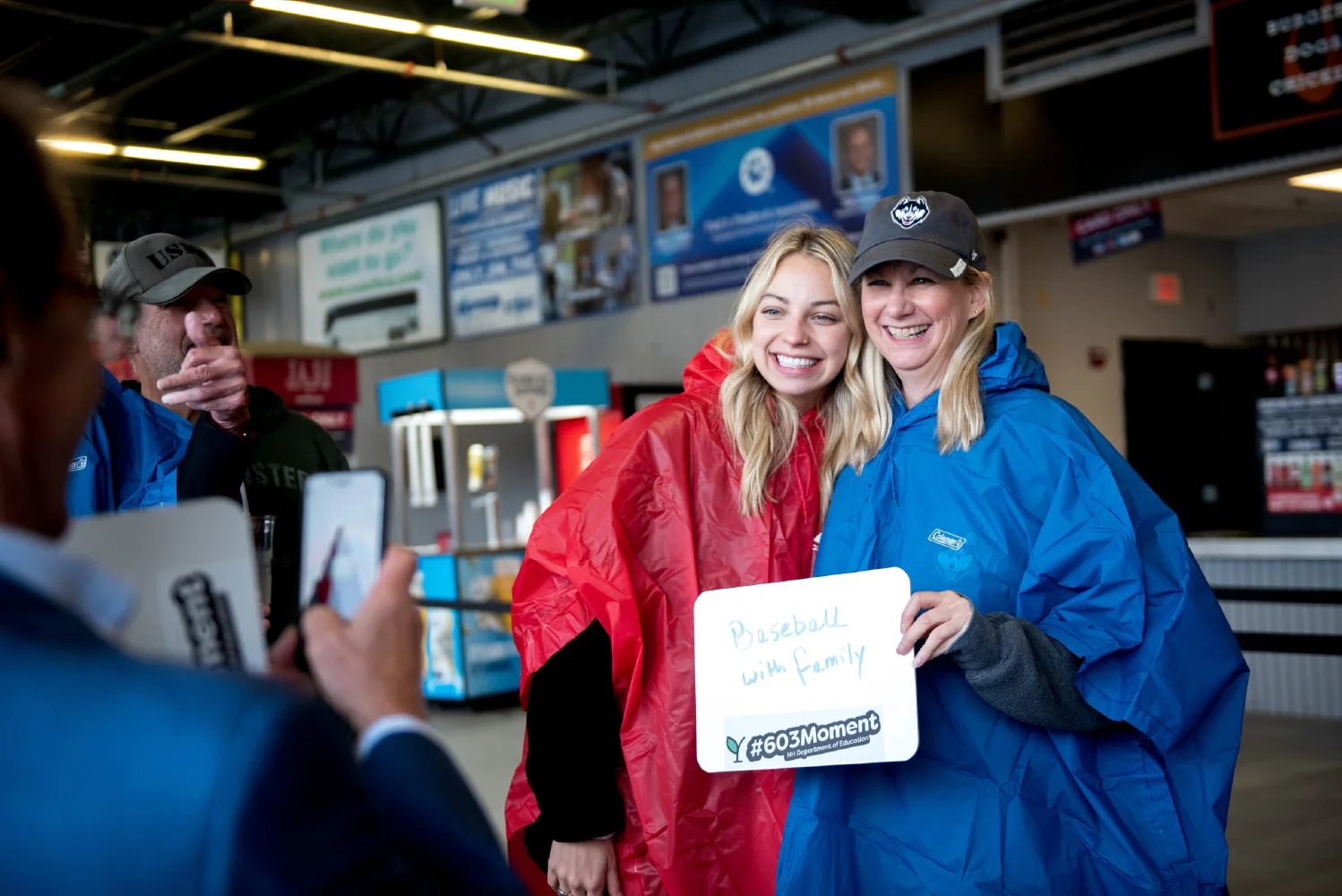Two women in rain ponchos smile for a photo, holding a "Baseball with family" sign—a picture-perfect moment that captures memories and the spirit of growth marketing by bringing communities together.