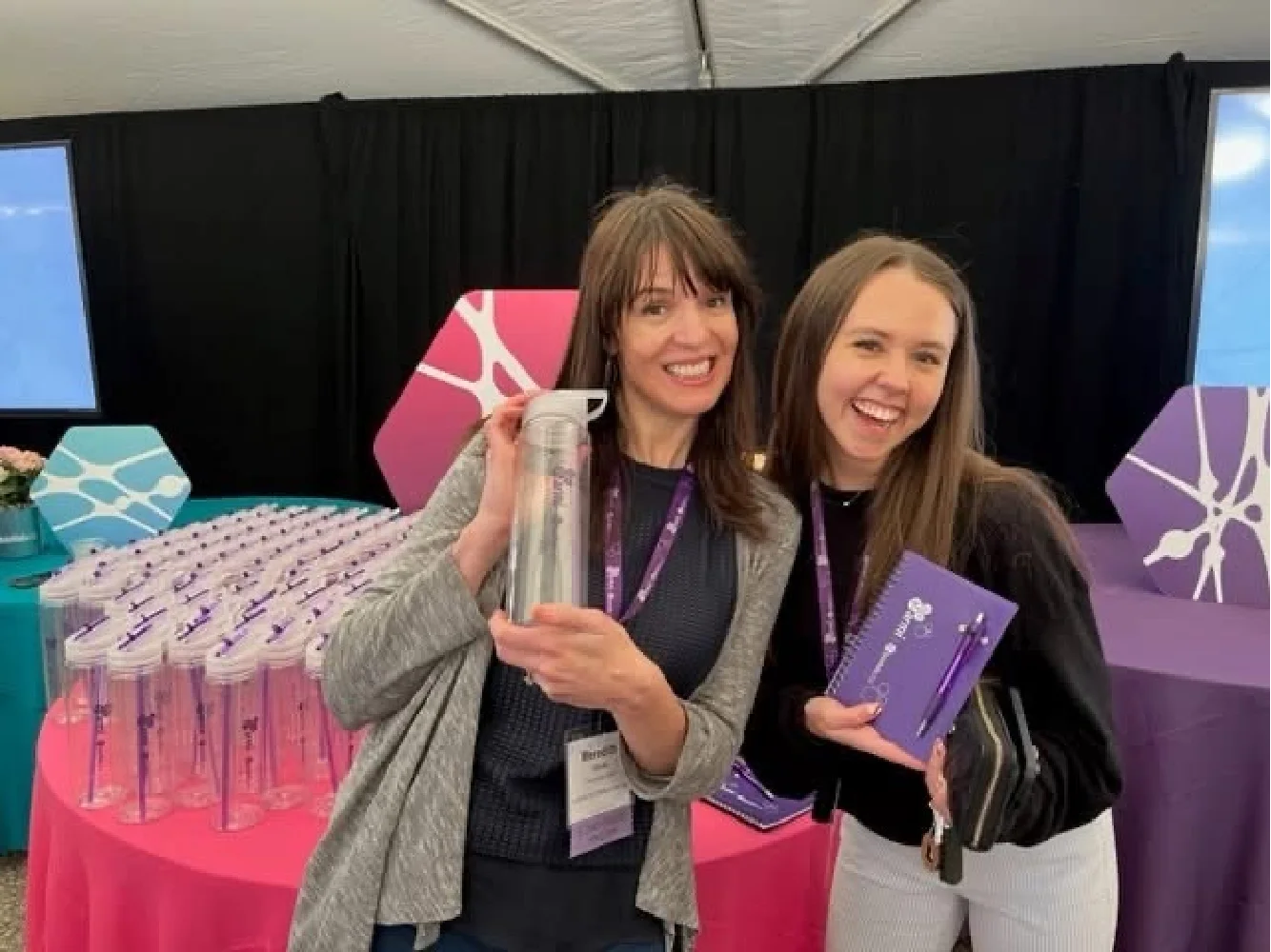 Two women smiling, holding growth marketing event swag items, with more gifts displayed on a table behind them.