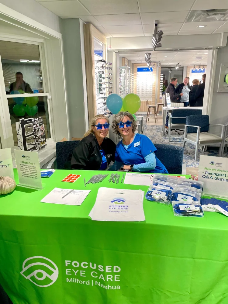 Two women in blue shirts sit at a Focused Eye Care booth with a green tablecloth and promotional items, sharing insights about growth marketing.
