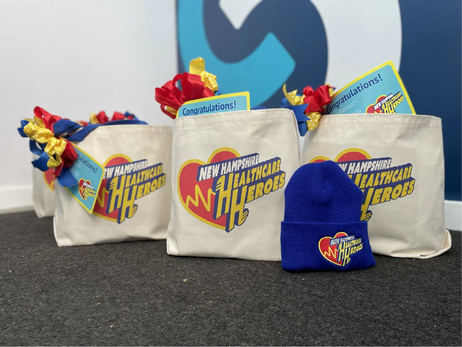 Canvas bags labeled “New Hampshire Healthcare Heroes” with gifts and a blue knit hat on a carpeted floor.