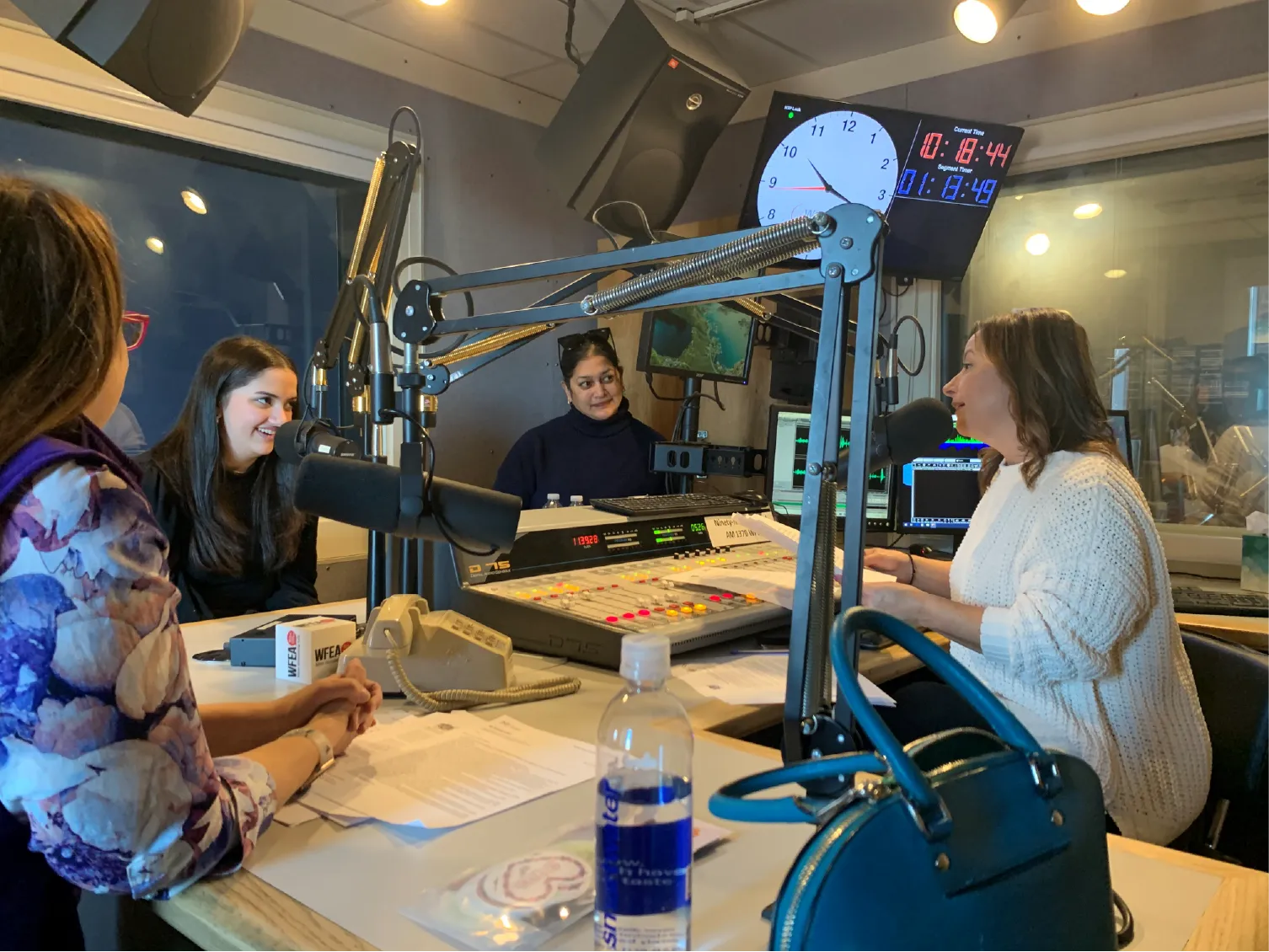 Four women sit around a table with microphones in a radio studio, engaged in conversation and smiling.