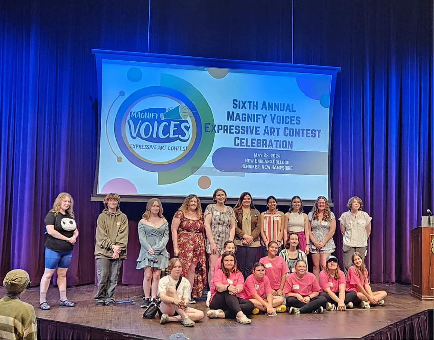 A group of people pose on stage under a sign for the Magnify Voices Expressive Art Contest Celebration.