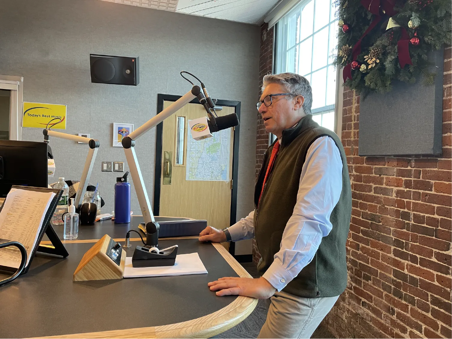 Man speaking into a microphone in a radio studio with brick walls and a large window with a wreath.