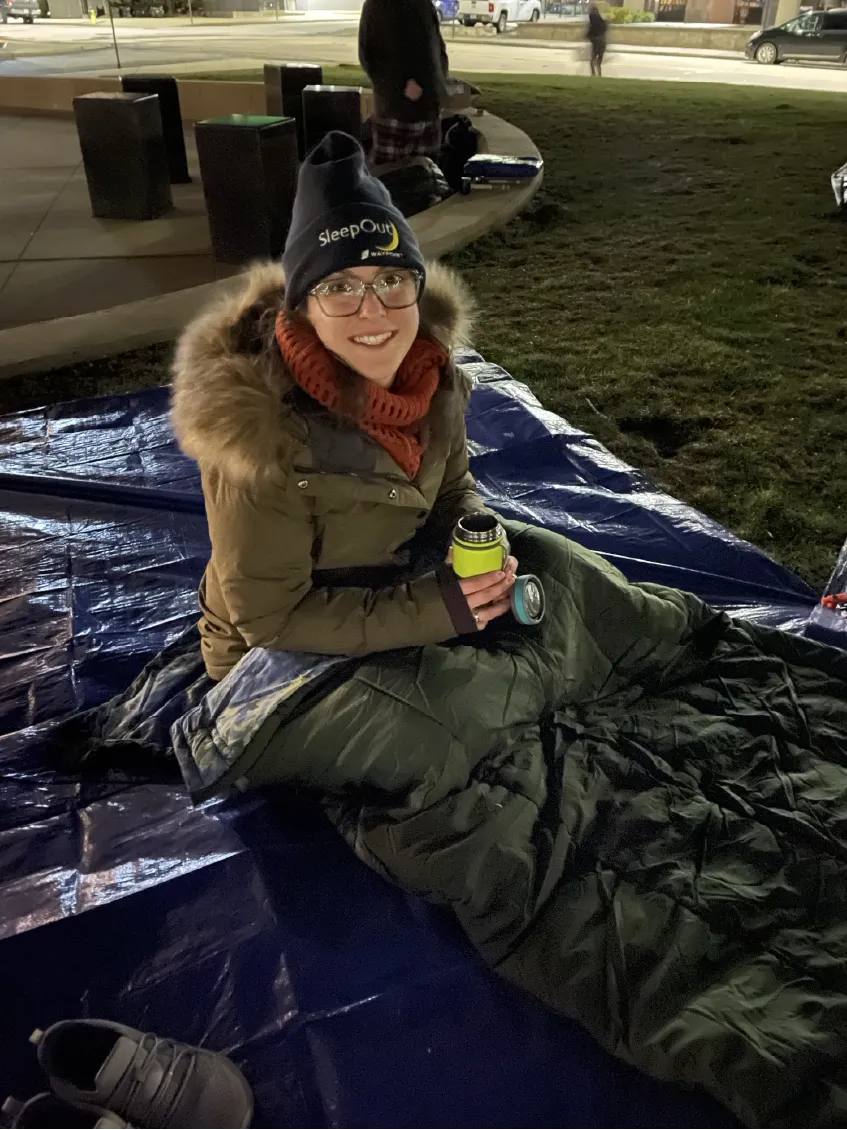 A person in a winter coat and hat sits in a sleeping bag on a tarp outdoors at night, smiling and holding a mug.