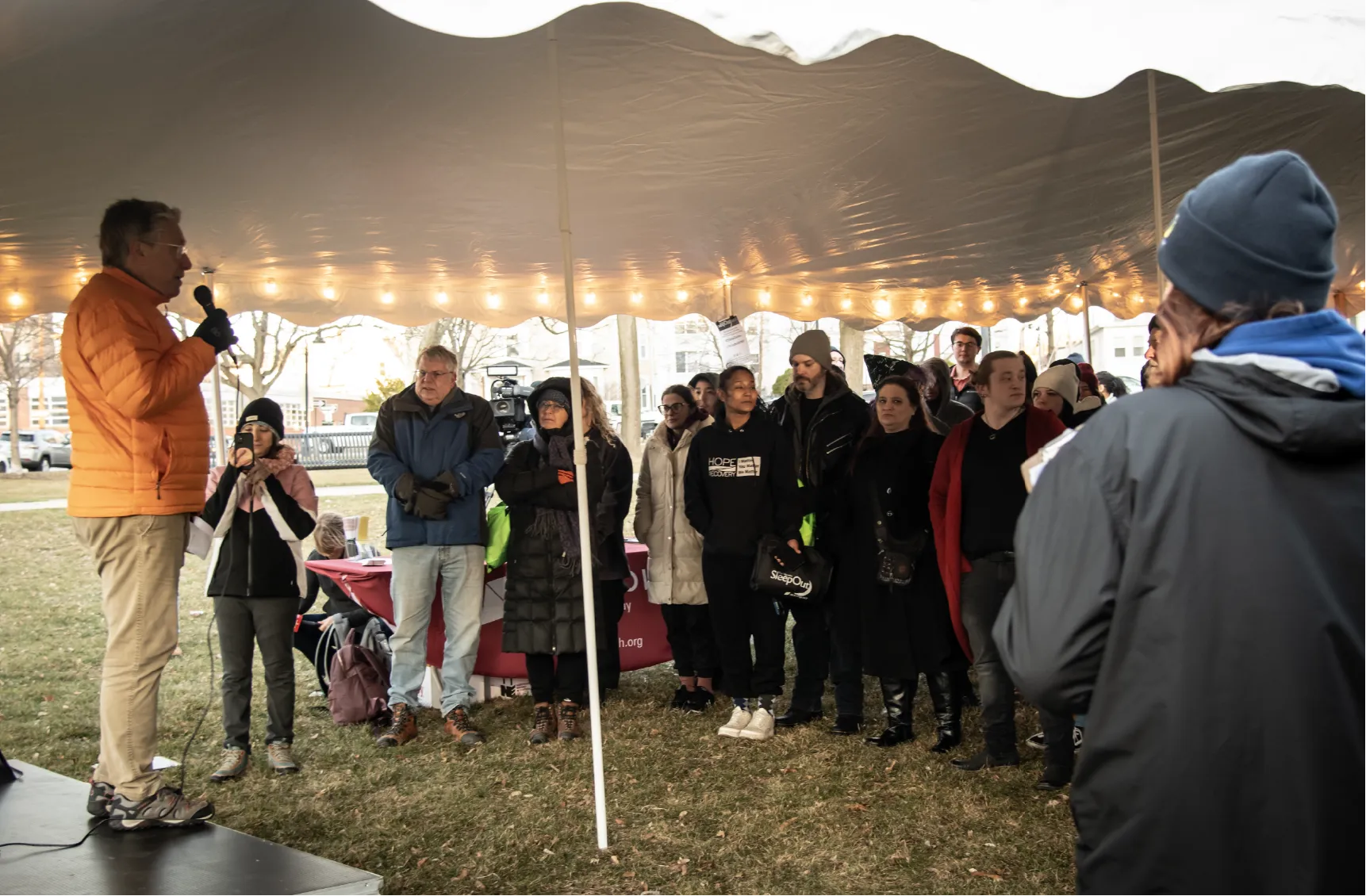 A speaker addresses a crowd under a tent at an outdoor event on a chilly day.
