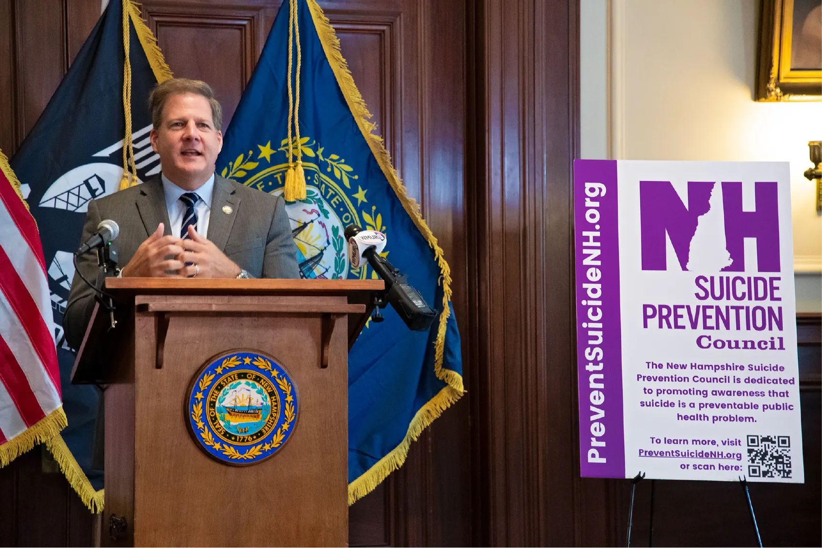 A man speaks at a podium next to a sign about the New Hampshire Suicide Prevention Council.