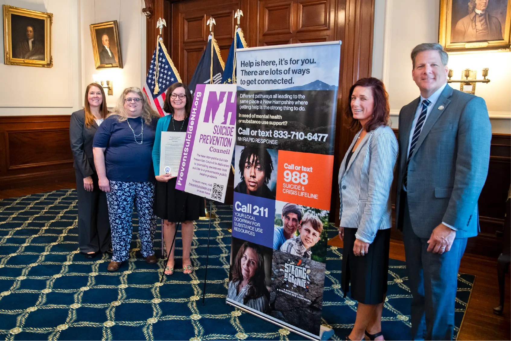 Six people stand indoors beside suicide prevention banners and U.S. flags, posing for a group photo.