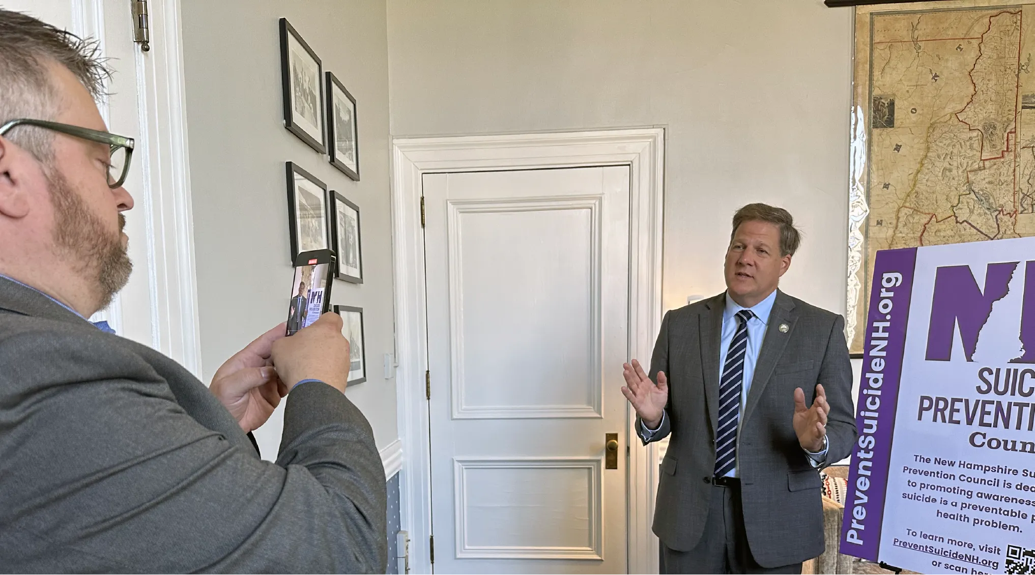 Two men in suits, one filming the other speaking near a suicide prevention poster in an office.