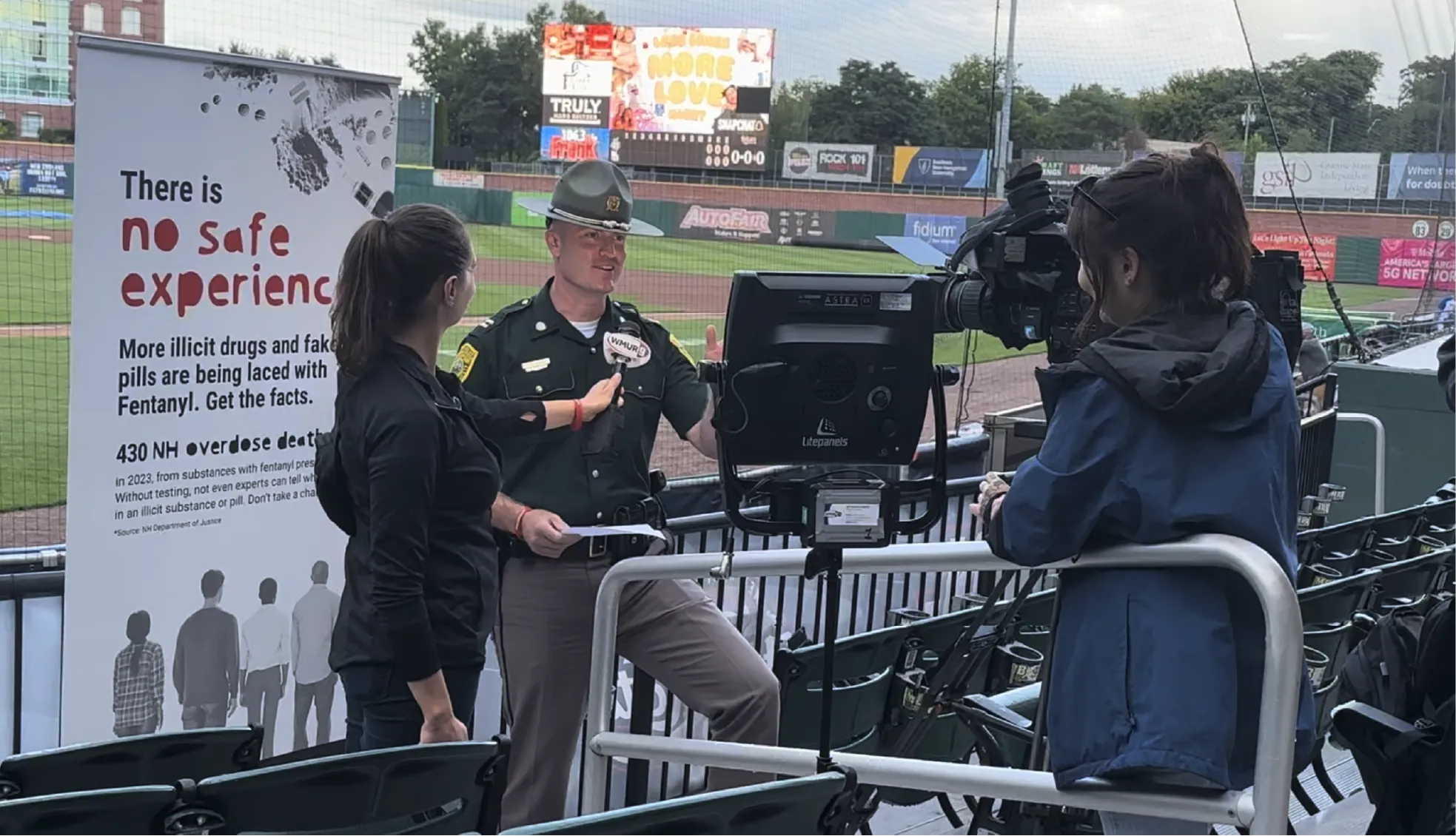 A police officer is interviewed on camera at a baseball stadium next to a drug safety awareness sign.