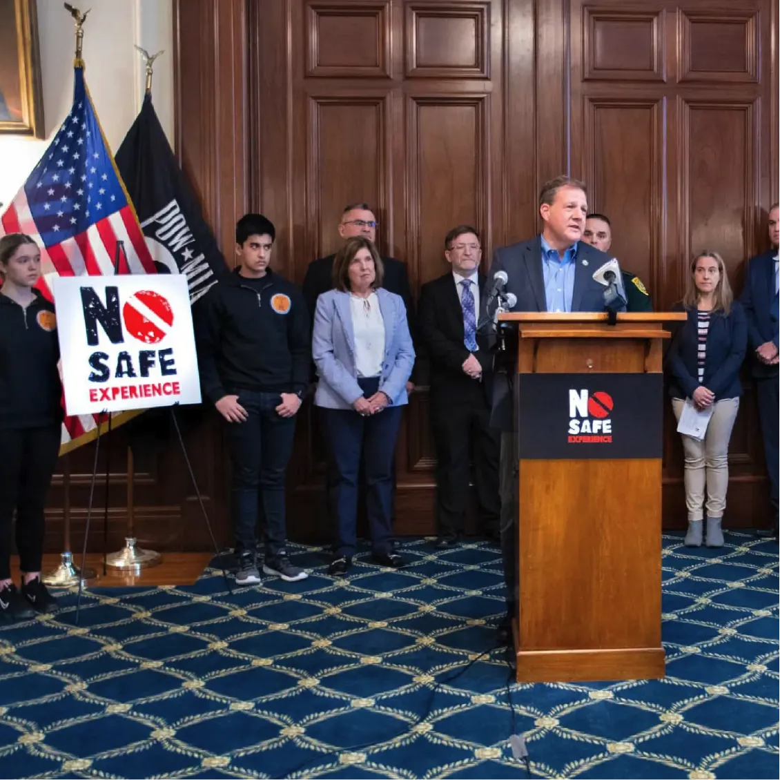 A man speaks at a podium with No Safe Experience signs, flanked by officials and uniformed youths.