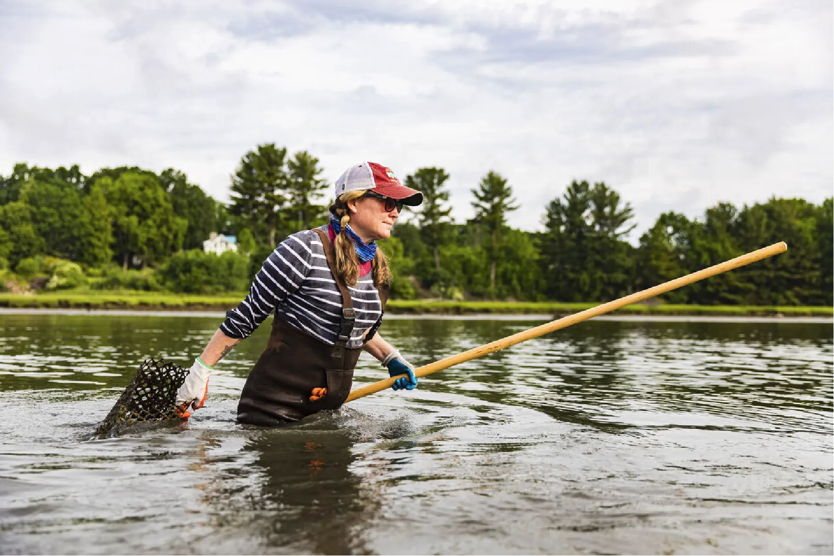 Person in waders stands in a river, holding a long wooden pole and a mesh basket, with trees in the background.