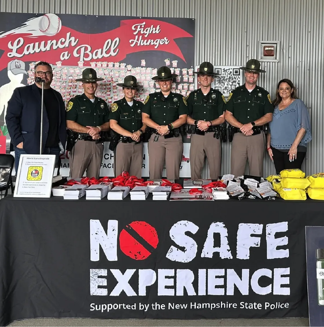 Seven people, including New Hampshire State Police officers, stand behind a table with No Safe Experience sign.