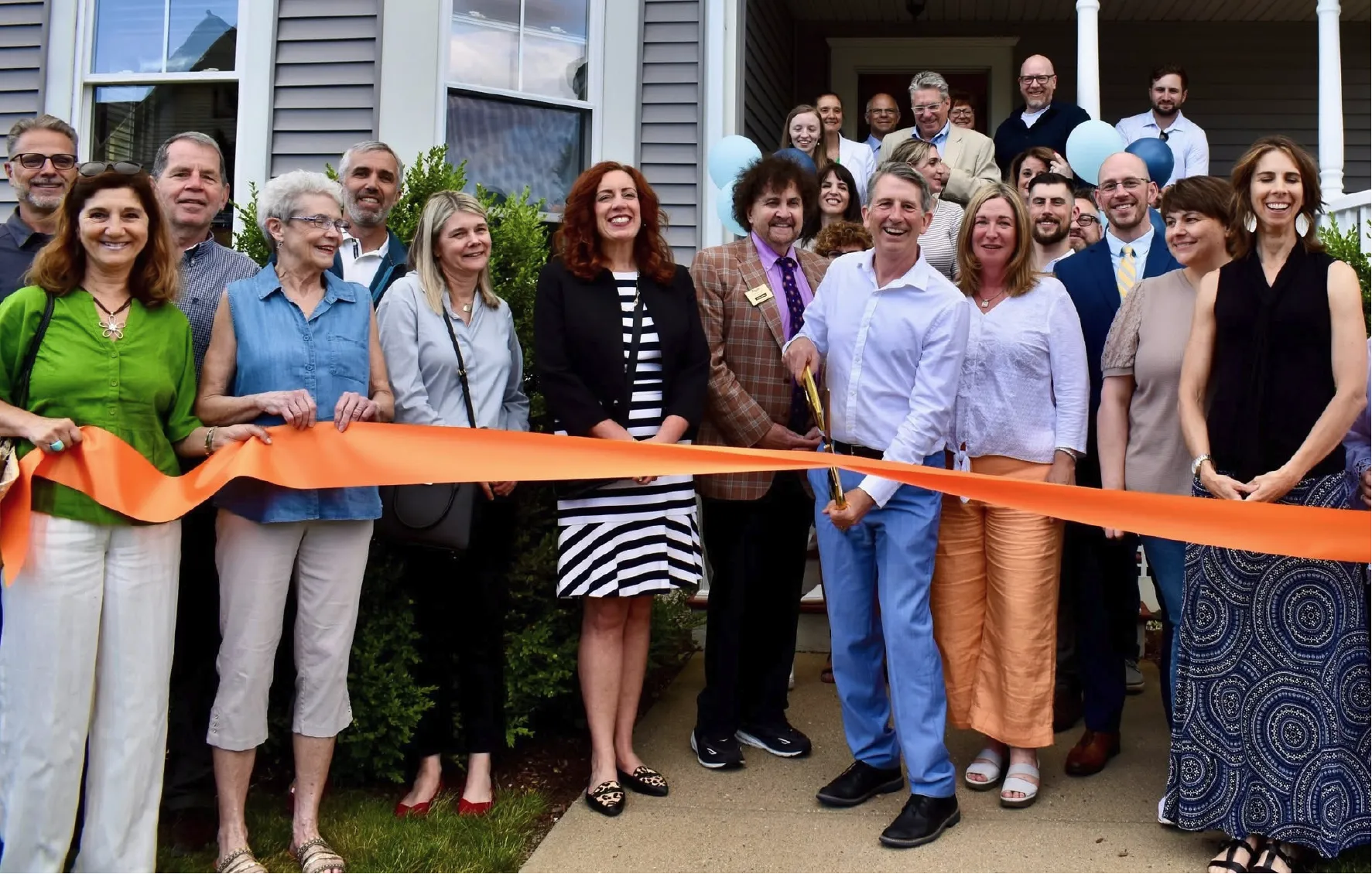 Group of people smiling at a ribbon-cutting ceremony in front of a house on a sunny day.