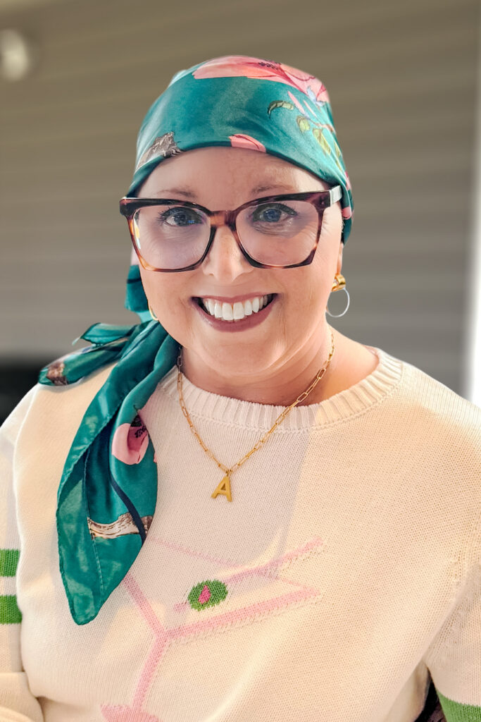 Smiling woman with glasses and a green floral headscarf, wearing a white sweater and gold A necklace.