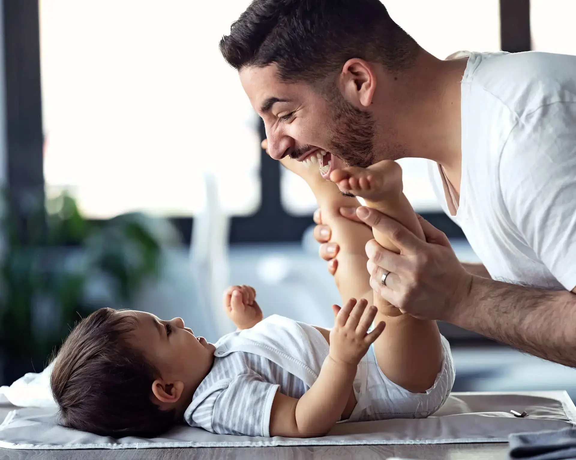 Smiling man playfully holds a baby’s legs as the baby lies on a changing table, both appearing happy.