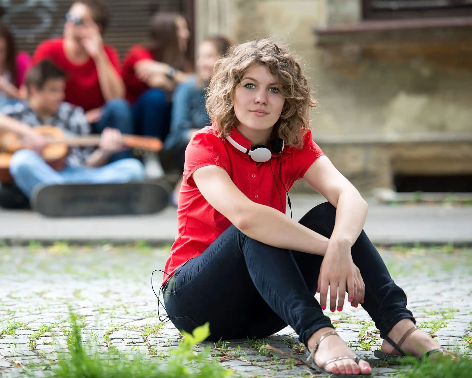 Young woman with headphones sits on cobblestones; friends in the background, some playing guitar.