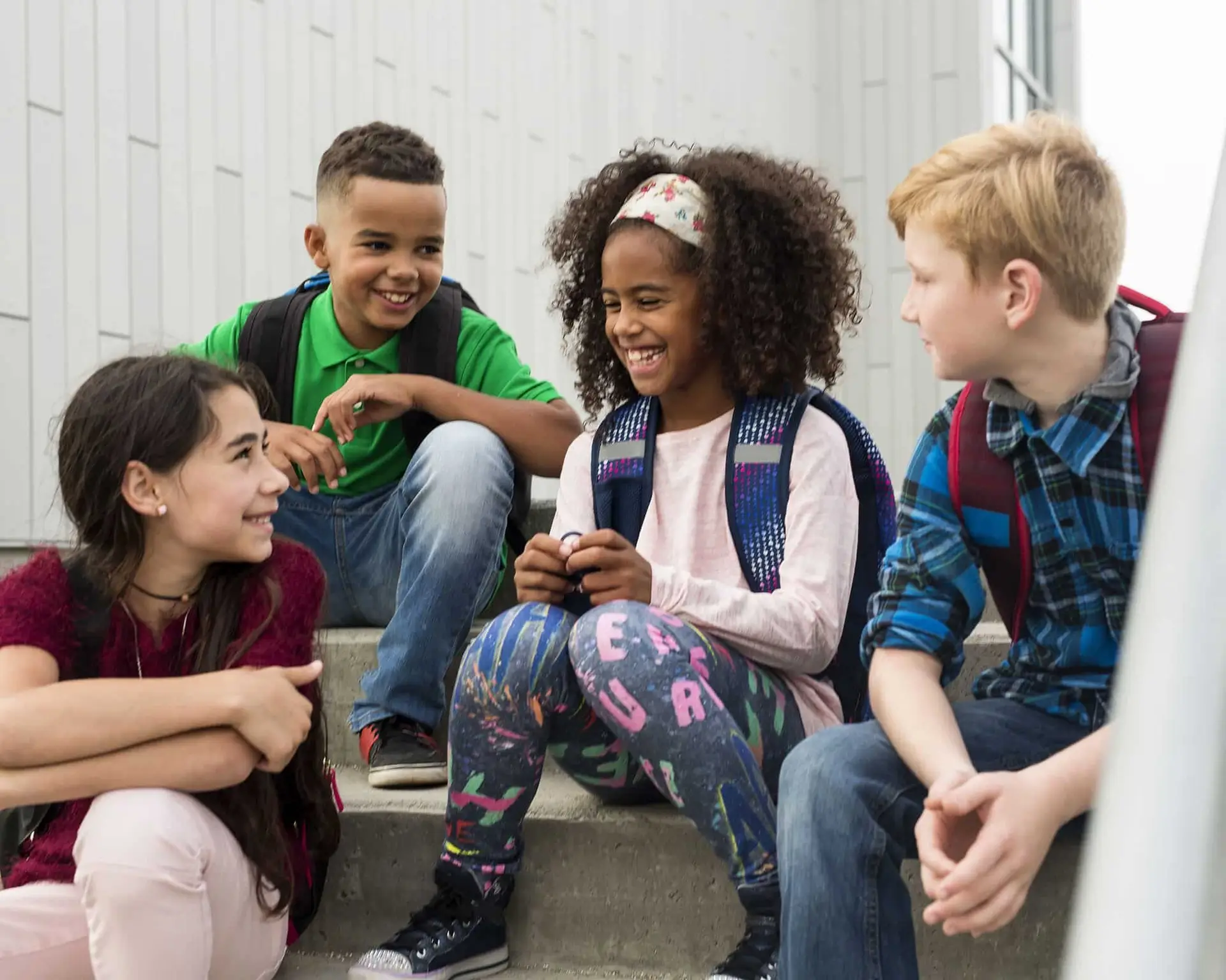 Four children with backpacks sit on outdoor steps, smiling and talking together.
