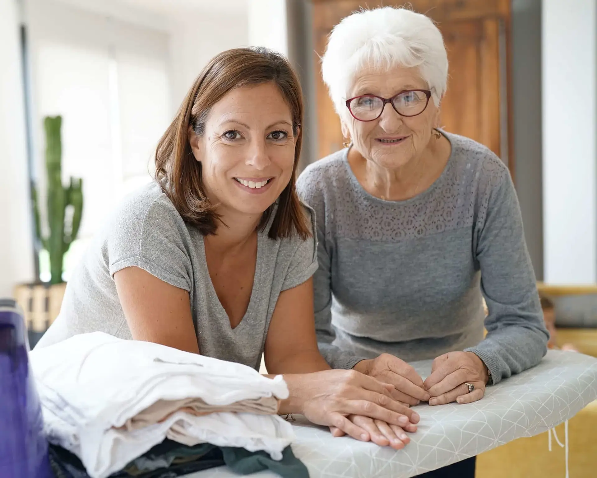 Two women, one younger and one older, smiling together while folding laundry on an ironing board indoors.